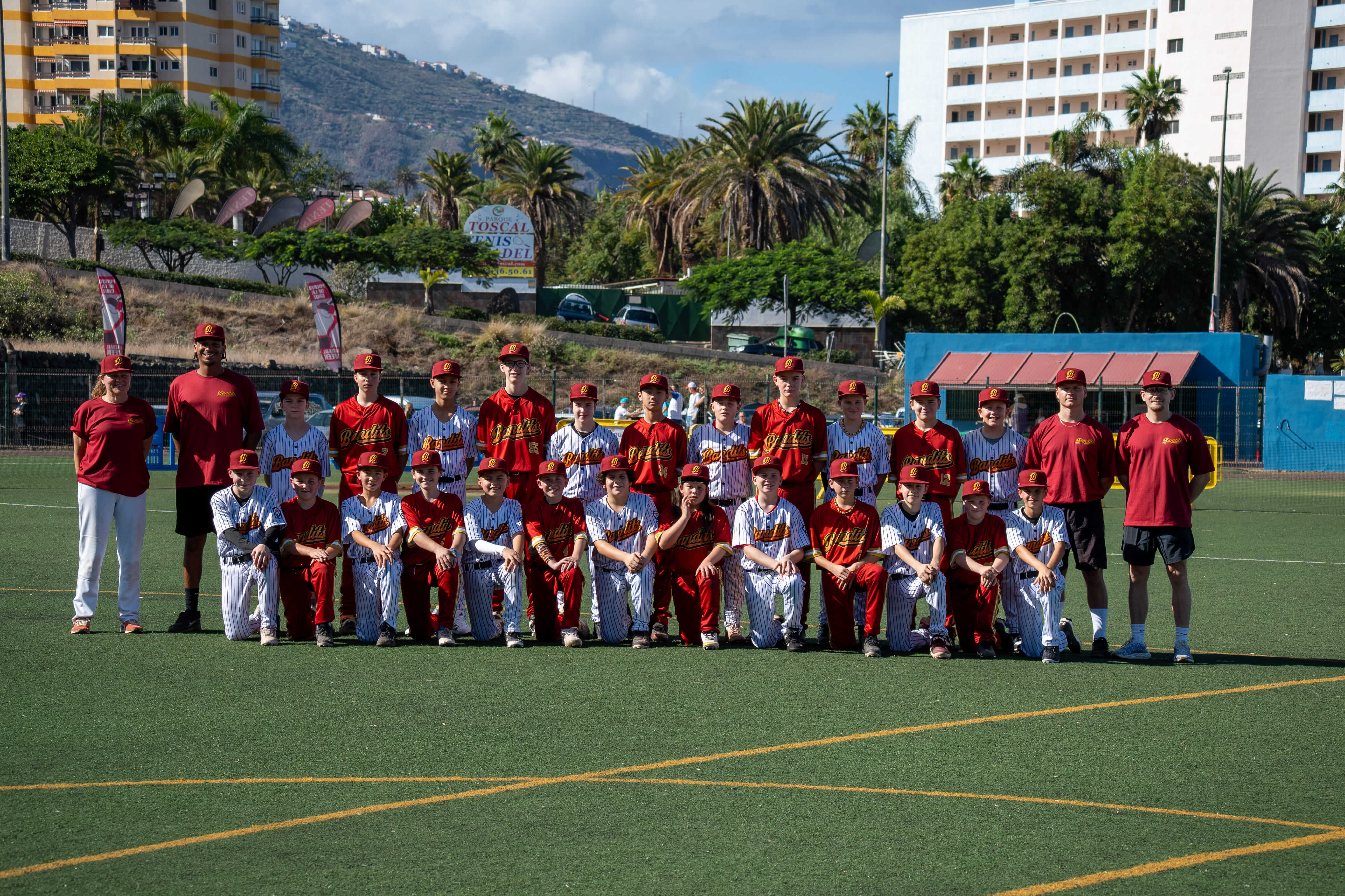 Zwei Jugend-Baseballteams der „Bandits“ in roten und weiß-gestreiften Trikots posieren gemeinsam mit mehreren Trainern auf einem Kunstrasenfeld. Im Hintergrund sind Palmen, Berge und Hotelgebäude bei sonnigem Wetter zu sehen.