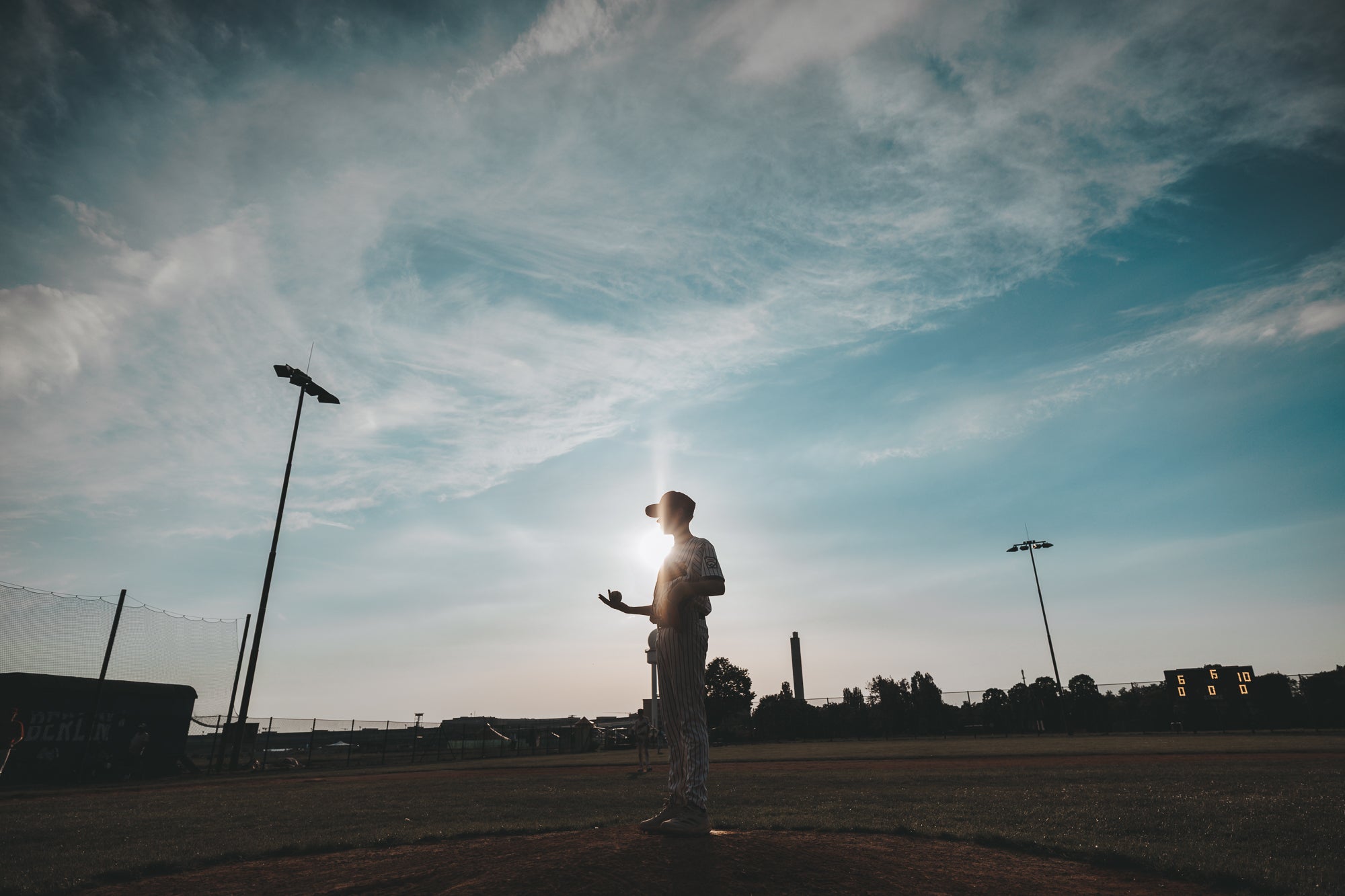 Silhouette eines Jugend-Baseballspielers auf dem Pitcher's Mound bei Sonnenuntergang während der Diamond City Series Berlin. Die tief stehende Sonne und der weite Himmel verleihen der Szene eine besondere Atmosphäre.