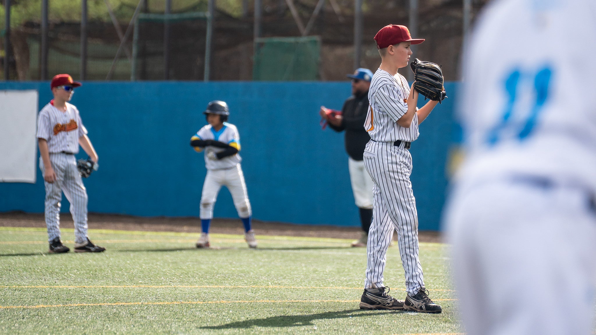 U13-Baseballspieler steht als Pitcher in der Wurfvorbereitung auf dem Feld, Mitspieler und Läufer im Hintergrund
